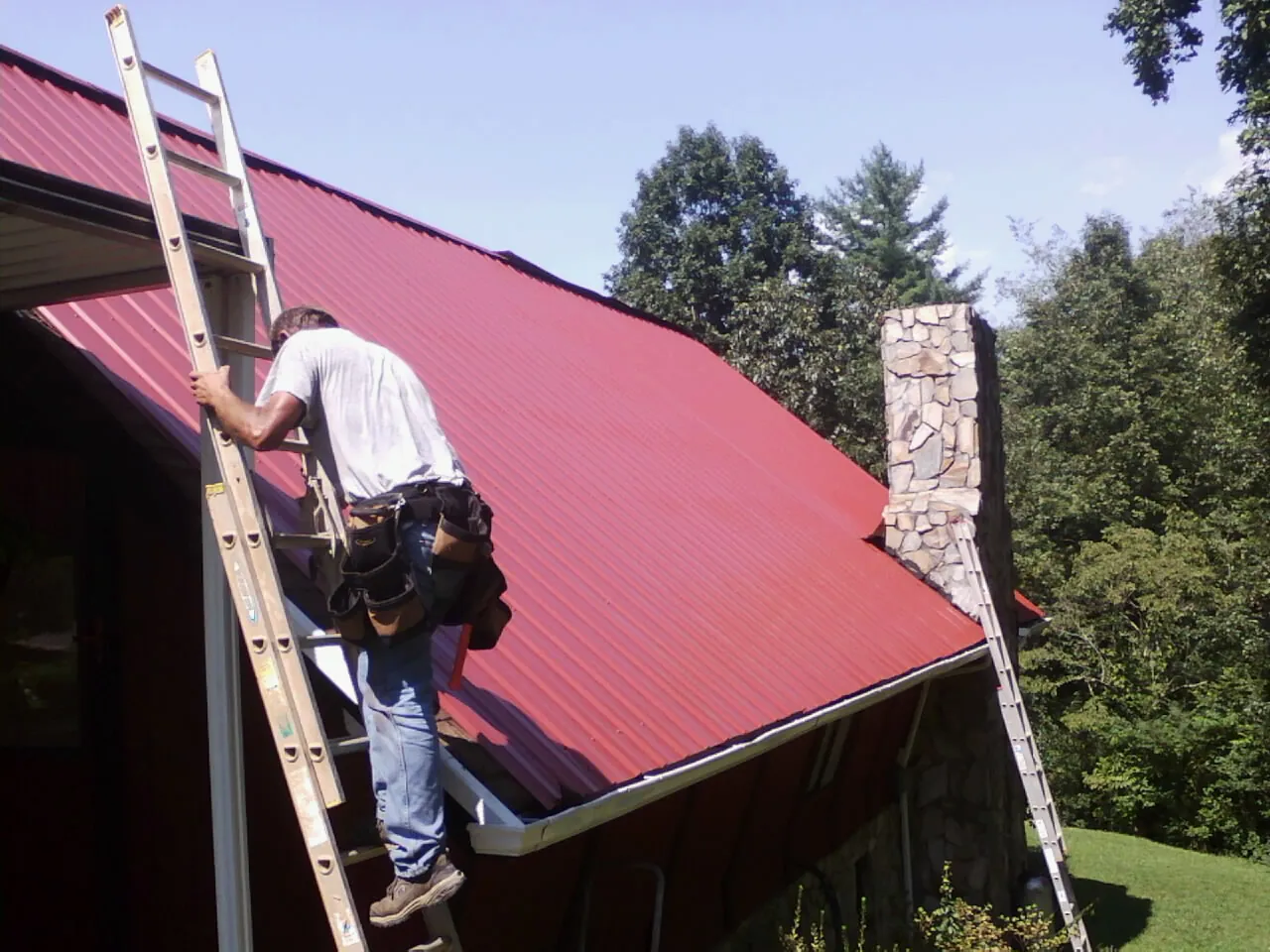 Roofer on a ladder beside a red metal roof and stone chimney — Soffit & Fascia Repair by Roofing Brothers in Seaside Park